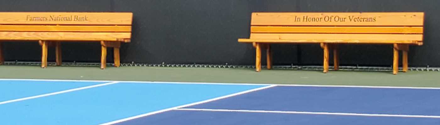 Wooden benches with memorial inscriptions by a tennis court.