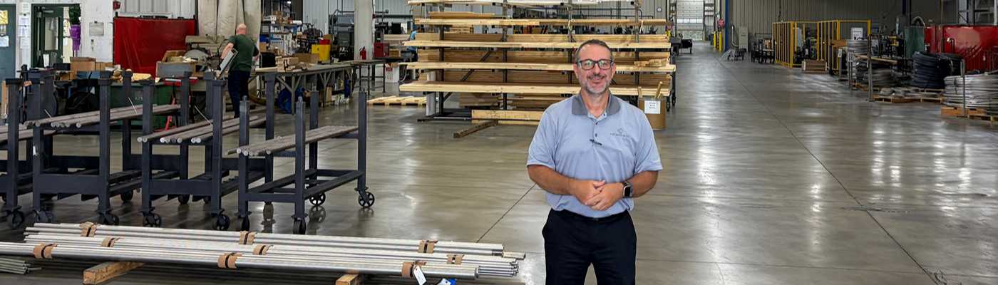 Man standing in a manufacturing facility surrounded by metal shelving and stock materials.